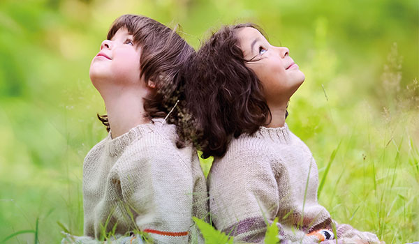 Two children sit back to back in the grass, looking happily up at the sky, both wearing bright knitted sweaters.